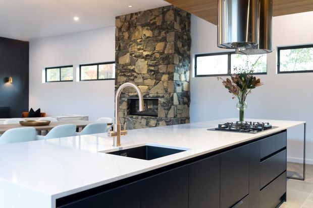 A bright contemporary open-plan kitchen featuring a large white island with an integrated sink, dark navy or black cabinetry, a gas cooktop, and a suspended stainless-steel rangehood. A substantial rough natural-stone fireplace or chimney structure forms a central feature behind the island, with a dining area and living space visible beyond. White walls, horizontal windows, recessed lighting, and timber ceiling detailing complete the interior.