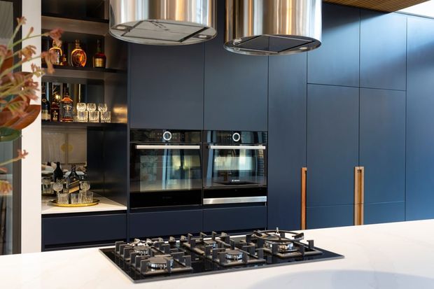 Contemporary kitchen featuring a large white island with an integrated black gas cooktop, deep navy-blue full-height cabinetry, timber-toned vertical handles, twin built-in ovens, and an open dark display and drinks cabinet. Two sculptural pendant rangehoods hang above the island, creating a refined, high-contrast scheme.