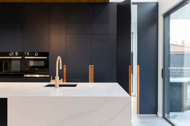 Contemporary kitchen featuring a large white stone island with integrated sink and brushed brass tap, dark matte full-height cabinetry with timber-toned vertical handles, and built-in black appliances. A glazed door and full-height window provide views to an exterior balcony or terrace.