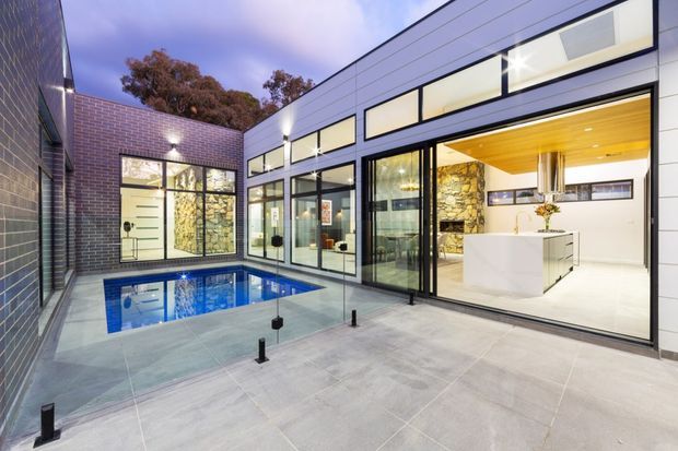 Contemporary courtyard-style home with a sheltered swimming pool and broad grey paved terrace. The building features dark brickwork, extensive black-framed glazing, a clean-lined white upper facade, and an open-plan kitchen visible through the glass, with a warm timber ceiling, white island, and exposed stone feature wall.