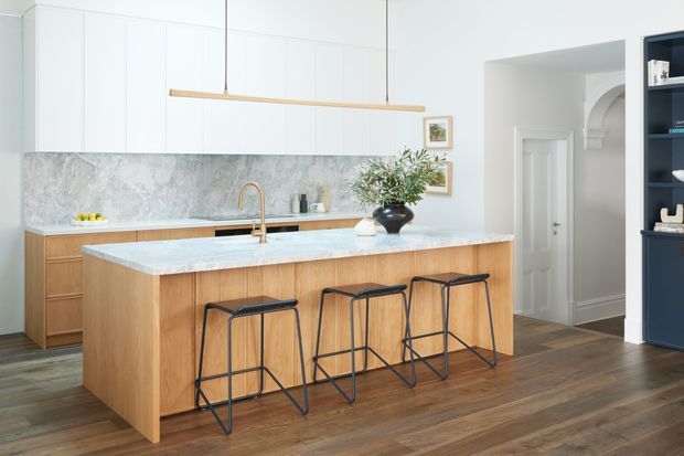 Bright, refined kitchen with a large timber island, pale stone benchtop, white full-height cabinetry, a textured stone-look splashback, and a long minimalist pendant light. The island includes seating for four on slim black stools, with dark timber flooring and a glimpse of an adjacent hallway and dark built-in shelving.
