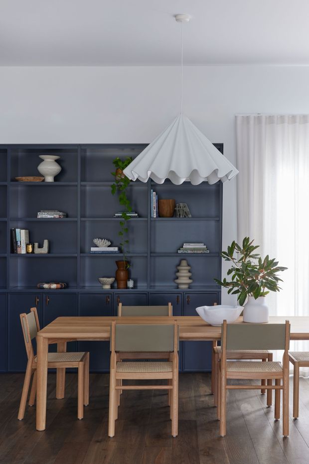 A refined dining room with a long natural timber dining table and matching woven-seat chairs, set against a full wall of dark navy built-in shelving and cabinetry. The shelves display books, ceramics, vessels and greenery. A sculptural white scalloped pendant light hangs above the table, while sheer curtains bring soft daylight into the room. Dark timber flooring and carefully styled decor create a warm, contemporary atmosphere.