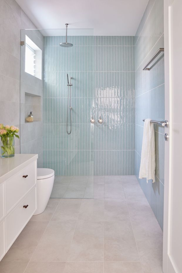 Bright contemporary bathroom with a walk-in glass shower, pale blue vertical wall tiles, large light grey floor tiles, white vanity cabinetry, concealed toilet, chrome fixtures and a white towel rail. Natural light enters through a high window, creating a clean and airy atmosphere.