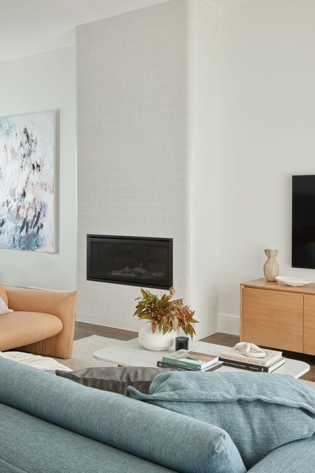 Bright, contemporary living room featuring a large white tiled fireplace surround with a black inset firebox, pale walls, a blue sectional sofa, blush-toned armchair, light timber media console, abstract artwork, and a sculptural coffee table with books and greenery.