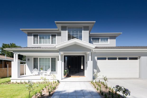 Front exterior view of a bright two-storey suburban house with a symmetrical gabled entry porch, white horizontal cladding, grey tiled or metal roofing, large windows, a covered veranda, landscaped front garden, and an attached double garage. The facade combines contemporary proportions with traditional residential detailing.