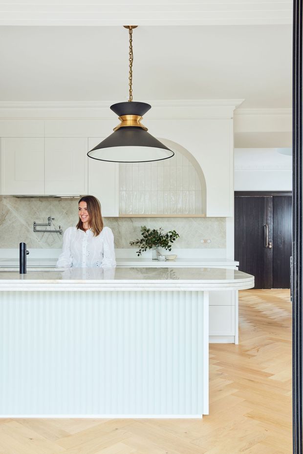 Bright, refined kitchen interior viewed from an adjoining space. The room features a large island with a pale fluted front and rounded stone benchtop, white shaker-style cabinetry, a pale marble or stone splashback, an arched tiled feature niche, brass-and-black pendant lighting, and light timber herringbone flooring. Dark fluted joinery frames the opening to the right, adding contrast to the predominantly white and warm neutral palette.