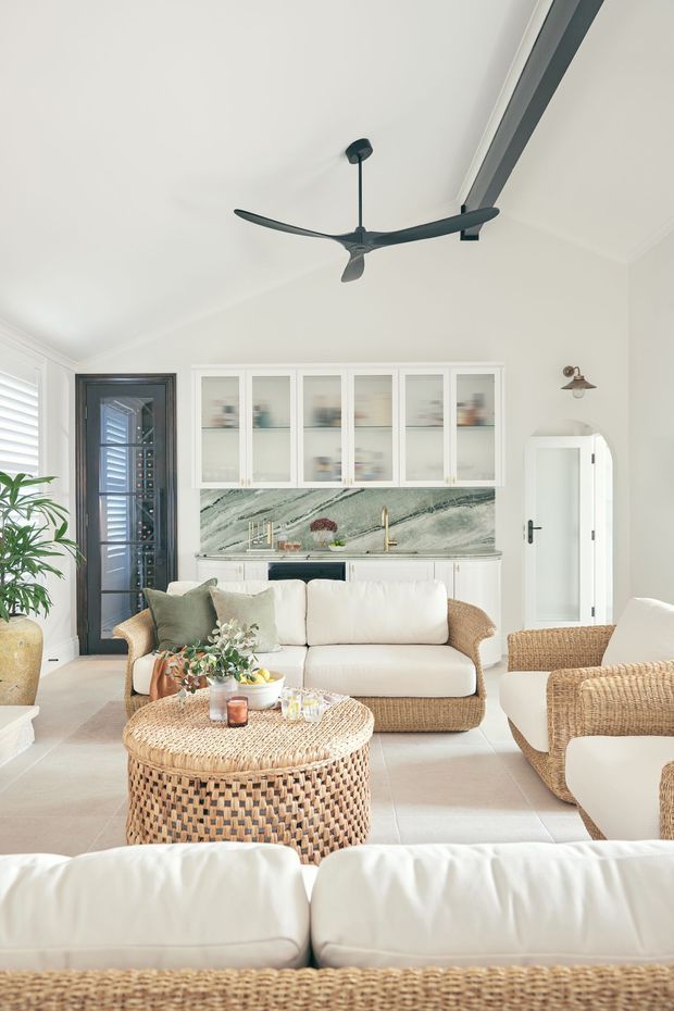 Bright, airy open-plan living room with a compact built-in kitchen at the rear. The room features white walls and cabinetry, a dramatic veined stone splashback, pale tiled flooring, woven rattan sofas and armchairs, a round woven coffee table, a dark ceiling fan, and a large linear skylight or ceiling glazing element. Shuttered glazing and a dark framed glazed door add architectural contrast.
