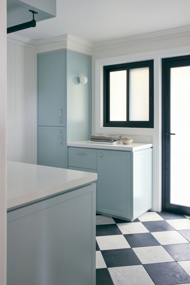 Bright, compact kitchen with pale blue-grey built-in cabinetry, a white benchtop, and a large island in the foreground. White vertical wall panelling and layered crown moulding create a refined traditional character, while black-framed glazing and a black-and-white checkerboard floor provide strong contrast. A small utility-style cabinet and counter sit beside the glazed external door.