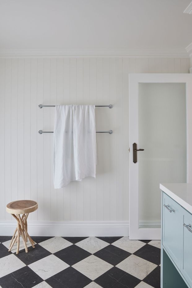 Bright bathroom interior with white vertical wall panelling, decorative cornice and skirting, chrome towel rails, a frosted-glass door, pale blue built-in vanity cabinetry, and a bold black-and-white diamond-pattern tiled floor. A small wooden stool adds a natural accent.