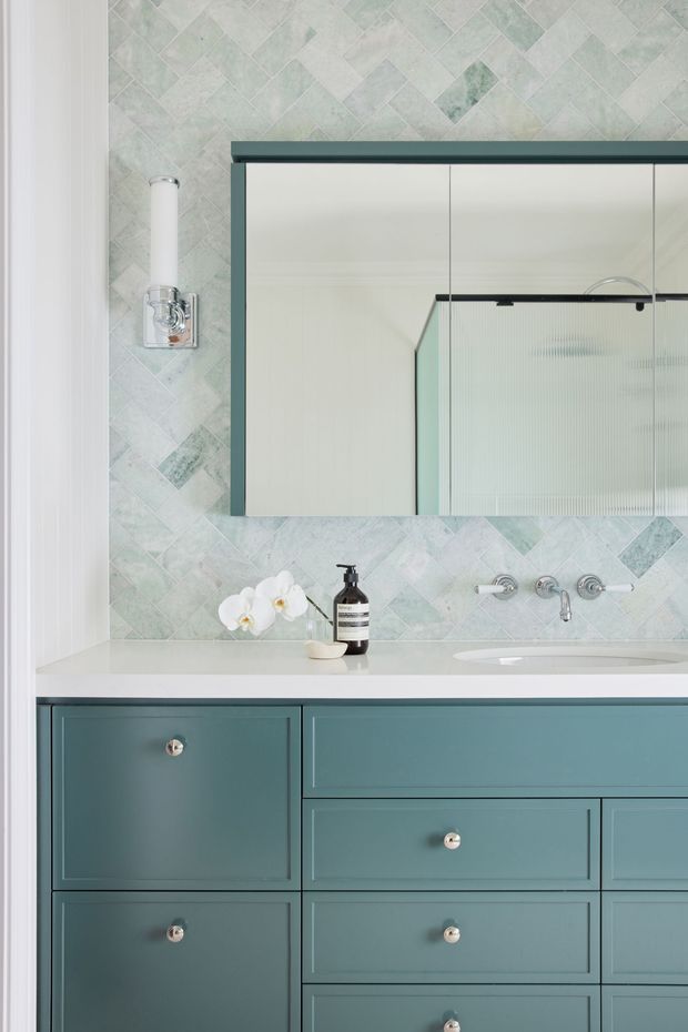 Close-up view of a refined bathroom vanity with muted blue-green shaker-style cabinetry, pale stone countertop, integrated basins and wall-mounted chrome fixtures. The wall is finished in pale green and white geometric patterned tile, with a large framed mirrored cabinet, fluted glass detail, and a traditional wall sconce. The palette is calm, cool, and softly elegant.