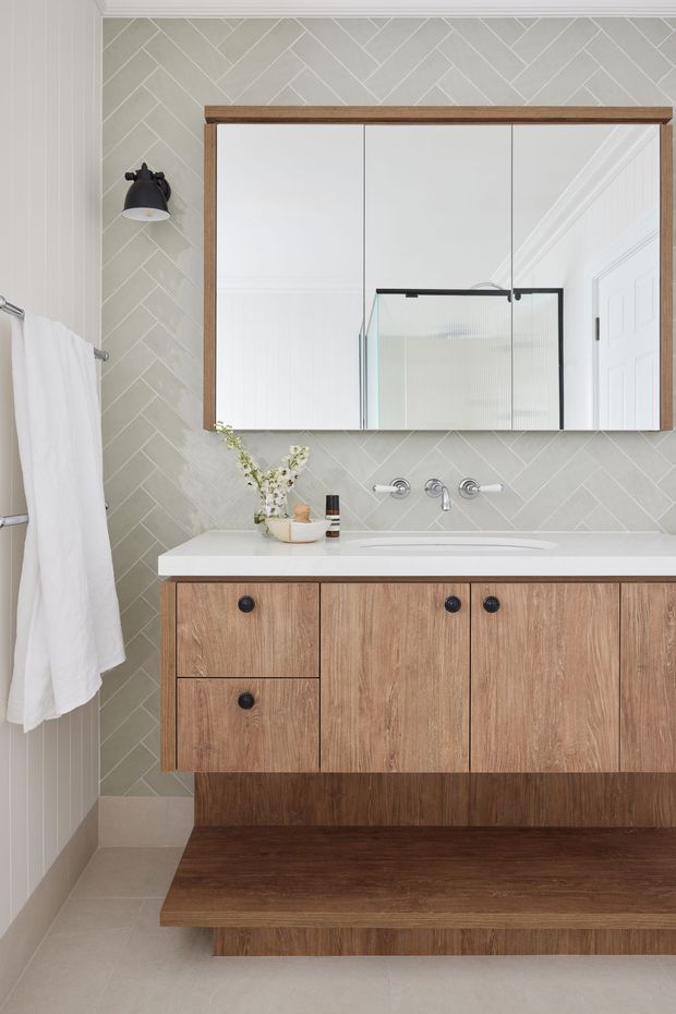 A refined contemporary bathroom vanity area with a floating timber-grain cabinet, white integrated basin, wall-mounted chrome tapware, and a large timber-framed mirrored cabinet. The walls are clad in pale grey-beige herringbone tiles, with white decorative trim, a black wall sconce, and a frameless glass shower enclosure reflected in the mirror. A white towel hangs on a side rail, and the dark timber platform below the vanity adds warmth and visual weight.