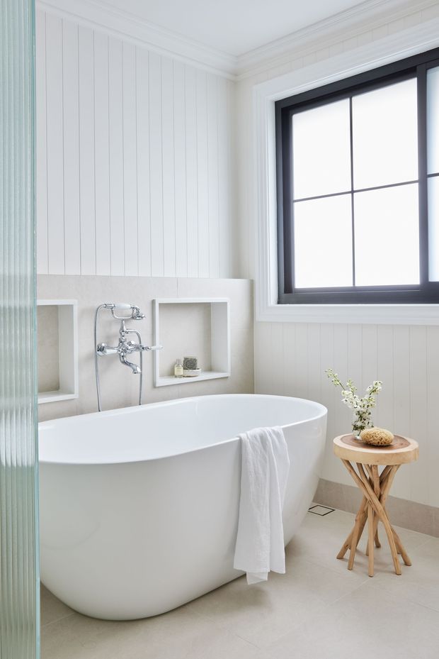 Bright, refined bathroom featuring a freestanding white bathtub, wall-mounted chrome bath fixtures, recessed display niches, white vertical wall panelling, pale stone-look tiled surfaces, and a large black-framed divided window. A small timber side table with simple botanical decor sits beside the bath, while a ribbed glass screen is visible at the left edge.