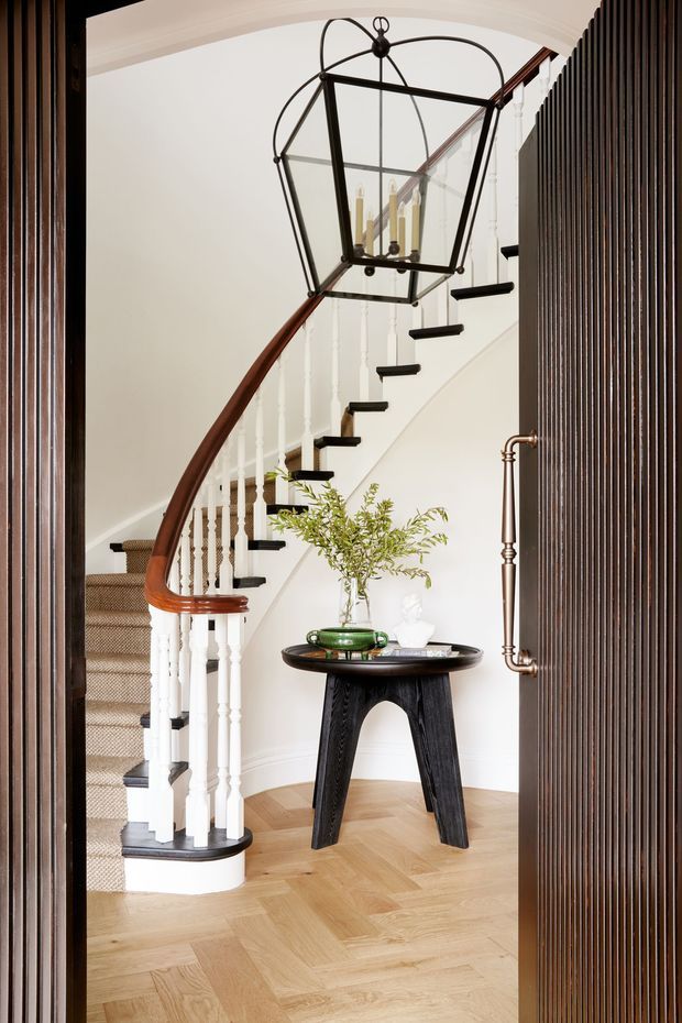 Elegant entry hall viewed through a dark fluted screen door, featuring a sweeping curved staircase with white turned balusters, dark stair treads, a warm timber handrail, and a large black metal-and-glass lantern pendant. A sculptural dark console table with a green vase and foliage sits on pale herringbone timber flooring. The space combines traditional detailing with contemporary decorative elements.