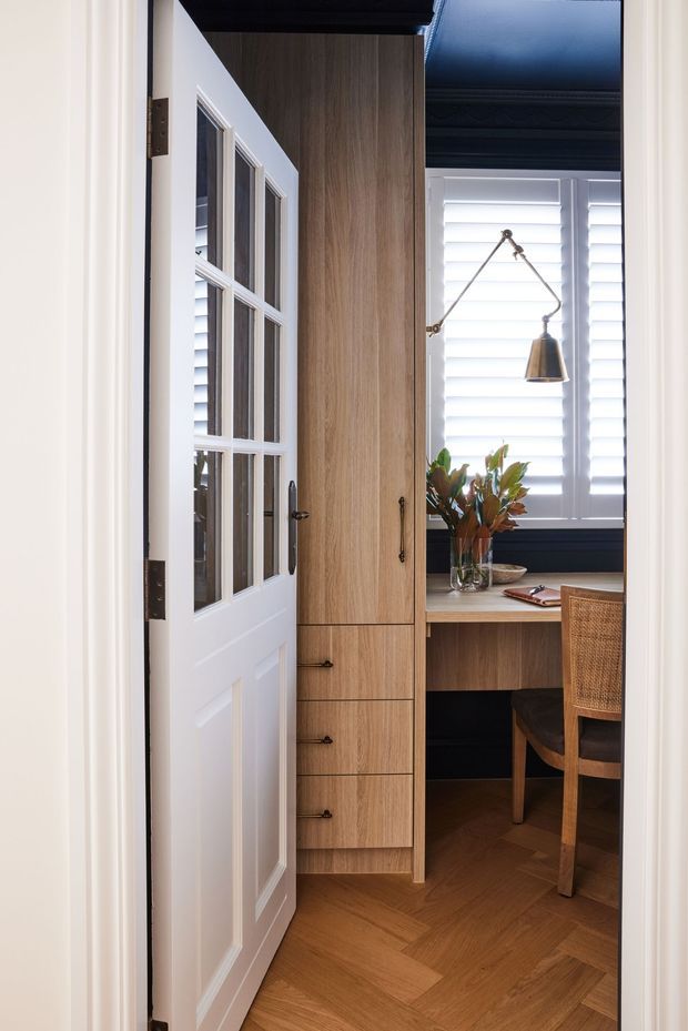 A compact, refined home office viewed through an open white-paned door. The room features custom mid-tone timber cabinetry with drawers and a built-in desk, a woven-seat chair, herringbone timber flooring, dark painted trim and ceiling detailing, white plantation shutters, and a brass articulated wall lamp. A small vase of foliage sits on the desk.