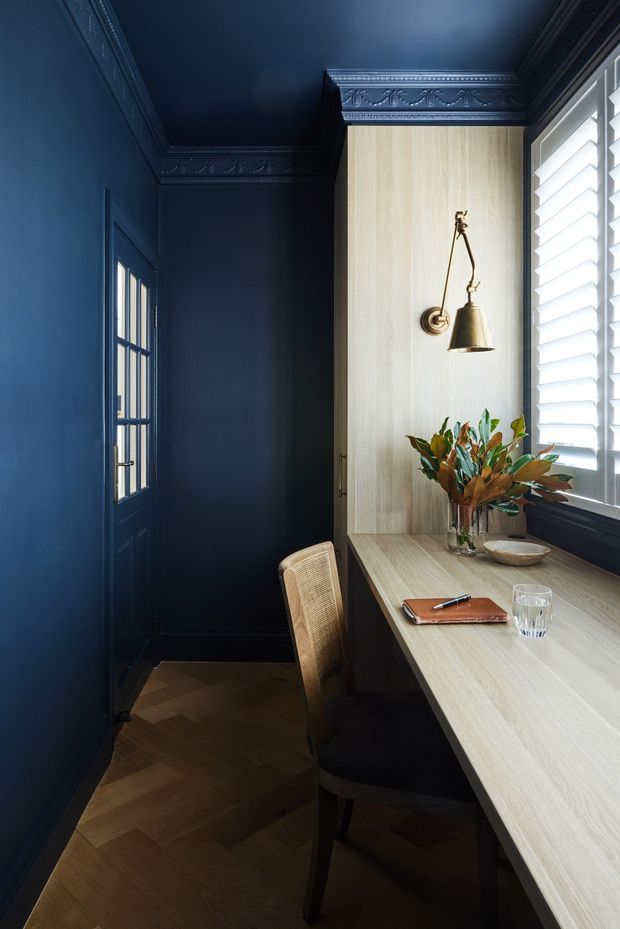 A narrow, richly detailed home office or study nook with deep navy-blue walls, ceiling, door, and ornate decorative cornices. A pale timber built-in desk runs beneath a large window fitted with white plantation shutters, with matching pale timber wall panelling behind a brass wall lamp. A woven-seat chair, notebook, glass, and vase of foliage sit on the desk. The herringbone timber floor and dramatic dark palette create a refined, intimate atmosphere.