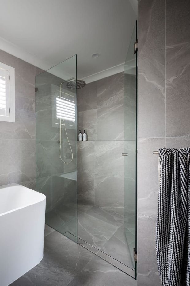 Contemporary bathroom featuring a frameless glass walk-in shower with a rainfall showerhead, large grey stone-look wall and floor tiles, a white freestanding bathtub, a small shuttered window, and a black-and-white towel on a wall-mounted rail. The room is bright, clean, and refined.