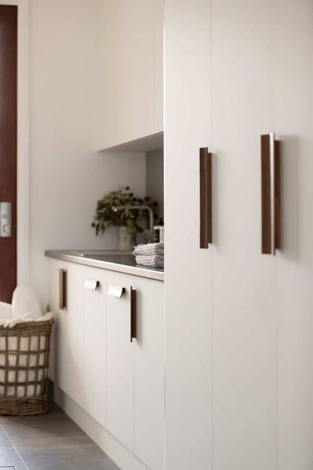 A compact, contemporary built-in laundry or utility area with full-height white cabinetry, dark timber handles, a dark benchtop, tiled flooring, and an integrated open shelf. A wicker laundry basket and folded towels reinforce the laundry function.