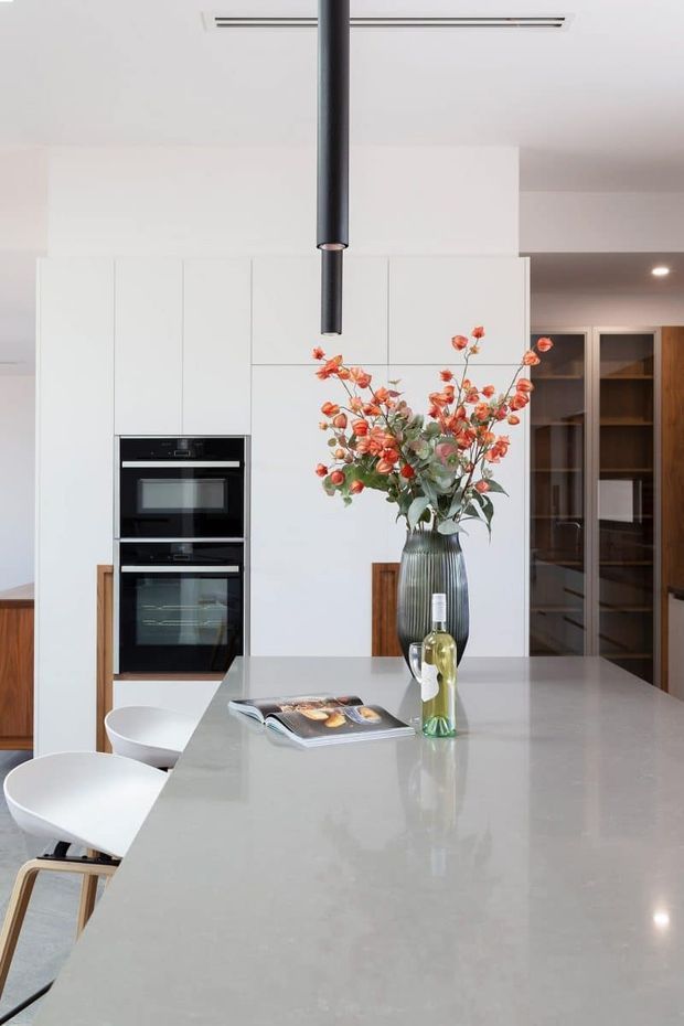 A bright contemporary kitchen centered on a large pale stone island with integrated seating. Full-height white cabinetry conceals two stacked black ovens, while a dark cylindrical rangehood hangs above the island. A tall ribbed vase with orange flowers and a bottle provide decorative accents. Timber details and a glazed display cabinet add warmth to the otherwise clean, minimalist space.