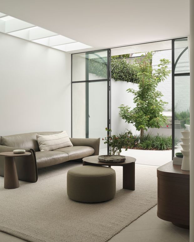 Refined contemporary living room with a low-profile grey sofa, rounded upholstered ottoman, dark wood side and coffee tables, and a neutral textured rug. Full-height black-framed glazing opens visually to a planted internal courtyard, while a linear skylight brings in abundant natural light. The space has clean white walls, restrained furnishings, and a calm indoor-outdoor connection.