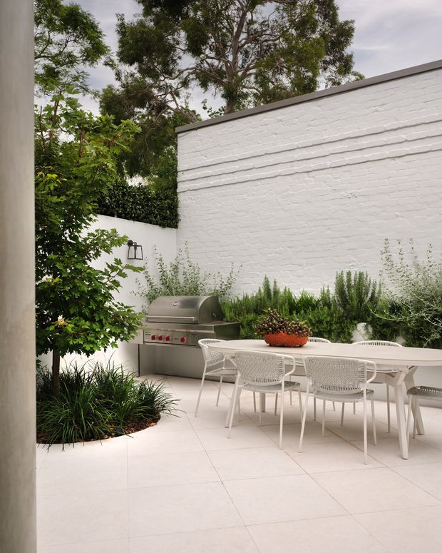 A refined outdoor dining courtyard with a white-painted brick privacy wall, pale tiled paving, white outdoor dining furniture, integrated stainless-steel barbecue, and dense green planting. The enclosed space has a clean, light contemporary aesthetic with a mature tree canopy visible above the walls.