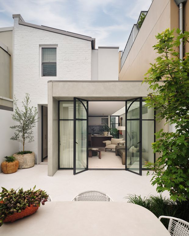 A contemporary courtyard patio viewed from an outdoor dining area, with pale rendered and white-painted brick walls, a concrete-framed opening, and black-framed glazed bi-fold doors leading into a refined living room. Potted trees and greenery soften the minimalist courtyard, while a round outdoor dining table and woven chairs occupy the foreground.