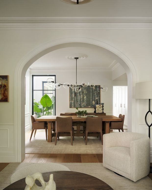 Elegant transitional interior viewed from a living area into a formal dining room through a large, heavily moulded arched opening. The dining room features a long dark timber dining table, upholstered brown dining chairs, a contemporary globe pendant, wall panelling, artwork, and a large rug. The foreground contains a light upholstered armchair and round timber coffee table. Neutral cream and white finishes, timber flooring, and abundant natural light create a refined, warm atmosphere.