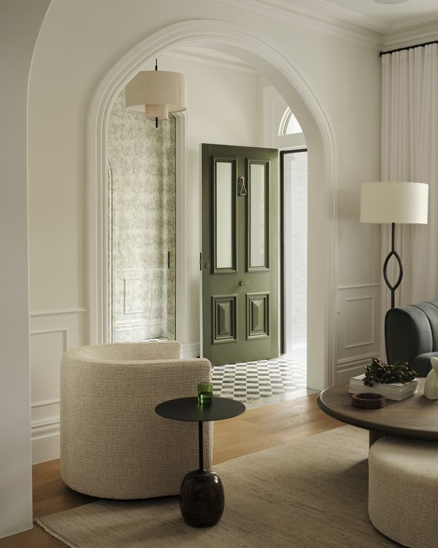 Refined residential entry and living area viewed from inside. The room features a large ornate arched opening with layered moulding, pale wall panelling, a dark green raised-panel front door, patterned checkerboard tile in the entry, warm timber flooring, and neutral upholstered seating. A black sculptural side table, round coffee table, soft curtains, and restrained contemporary furnishings create an elegant, calm transitional interior.