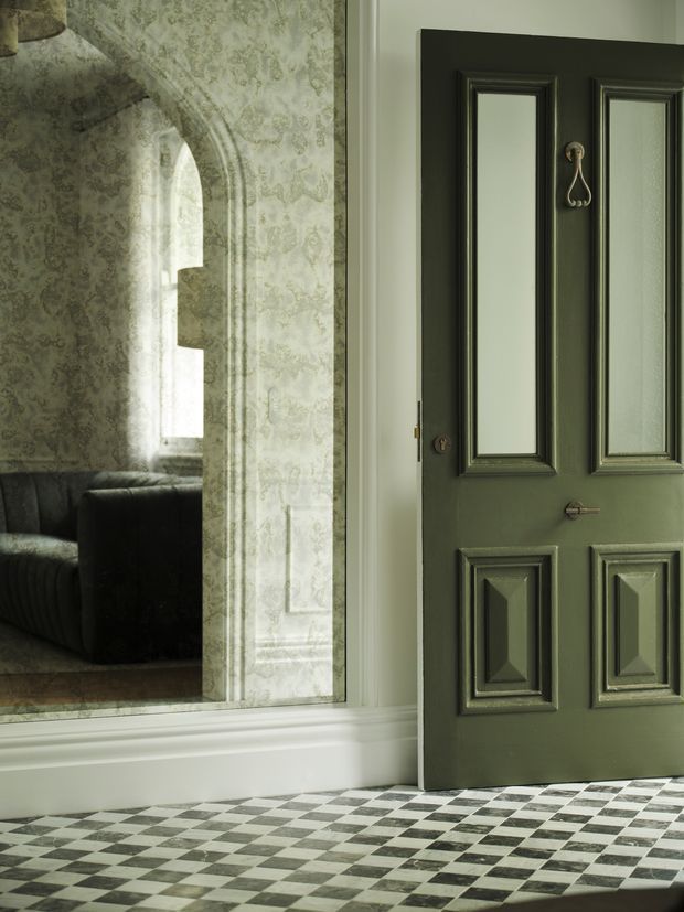 A refined interior view featuring a dark green, raised-panel door with decorative hardware, opening toward a sitting room visible through an ornate arched opening. The walls have pale patterned wallpaper and detailed white moulding, while the foreground floor is finished in a striking black-and-white checkerboard pattern. The overall composition has a heritage-inspired, elegant character with muted green, cream, and charcoal tones.