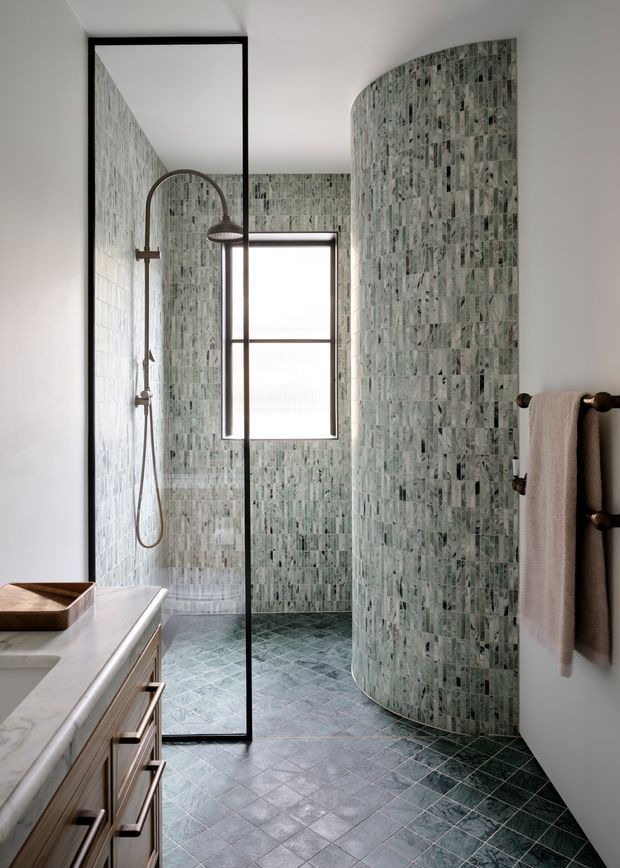 A refined contemporary bathroom featuring a walk-in shower enclosed by a black-framed glass partition, a curved tiled shower wall, and a floating-style timber vanity with a pale stone benchtop. Green-grey vertically patterned wall tiles continue onto the floor in a diagonal layout. Bronze-toned shower fittings and towel hardware add warmth, while a frosted window provides natural light.