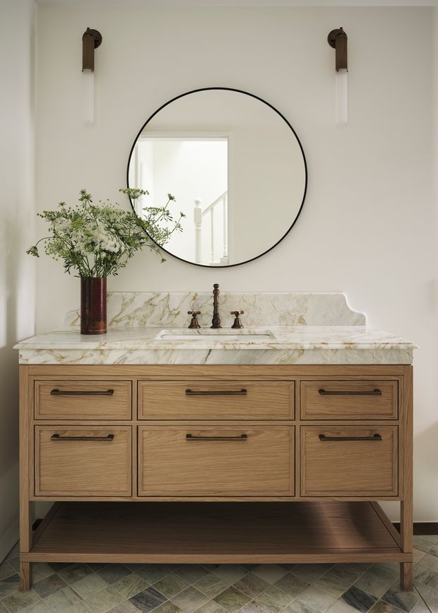 A refined bathroom vanity vignette featuring a freestanding timber vanity with open lower shelving, six inset drawers and dark metal pulls. The vanity has a white veined marble benchtop and integrated raised backsplash, paired with aged bronze or dark metal tapware. A large round black-framed mirror, matching wall sconces, and a vase of branches sit against warm white walls. Patterned stone or tile flooring is visible below.