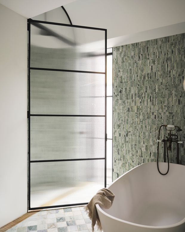 Contemporary bathroom featuring a freestanding white oval bathtub, a dark metal-framed reeded glass screen, a wall-mounted shower with aged dark-metal fittings, and a full-height green-grey patterned stone or marble tile feature wall. The floor is finished with small pale patterned tiles, and natural light enters through the translucent screen.