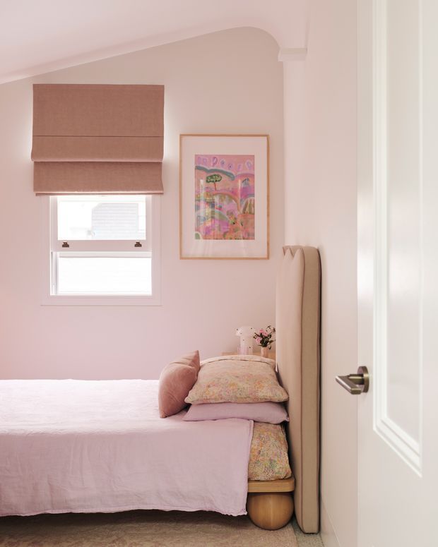 A compact, softly styled pink bedroom viewed through a partially open white paneled door. The room features a single bed with blush pink bedding, a tall upholstered beige headboard, patterned cushions, a small bedside surface, a Roman blind over the window, and colourful framed artwork. Traditional curved ceiling detailing and warm, muted tones create a cosy atmosphere.