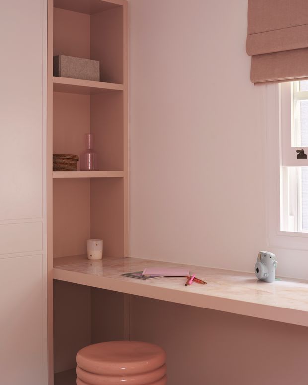 A compact built-in home workspace in a monochromatic dusty-pink scheme. The desk has integrated open shelving and cabinetry, a pale stone-like or painted worktop, and a rounded pink stool. A small window with a Roman blind provides natural light, while minimal stationery and decorative objects sit on the desk and shelves.
