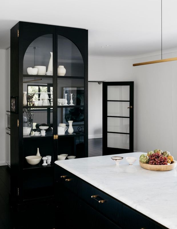A refined open-plan kitchen and dining area featuring a large black island with a pale stone benchtop, a tall black glass-fronted display cabinet, and matching black-framed internal doors. White walls and ceilings create a bright, high-contrast interior, with brass hardware and a linear pendant light adding warmth.