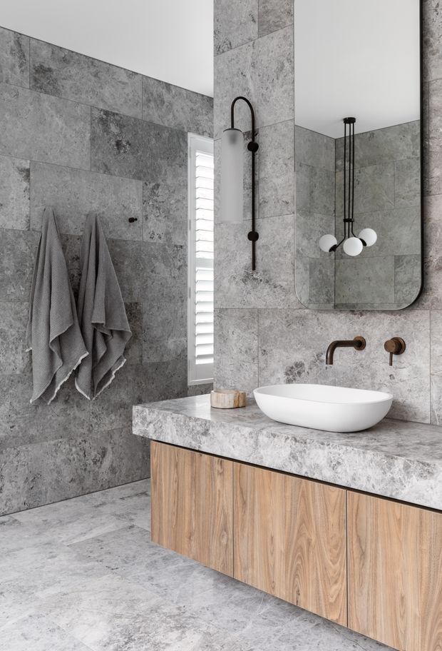A contemporary bathroom clad in large grey stone-look tiles, featuring a floating timber vanity with a thick stone benchtop, white vessel basin, wall-mounted bronze tapware, and a frameless rounded mirror. A vertical louvred window provides natural light, while black sculptural shower hardware and pendant lighting add contrast.