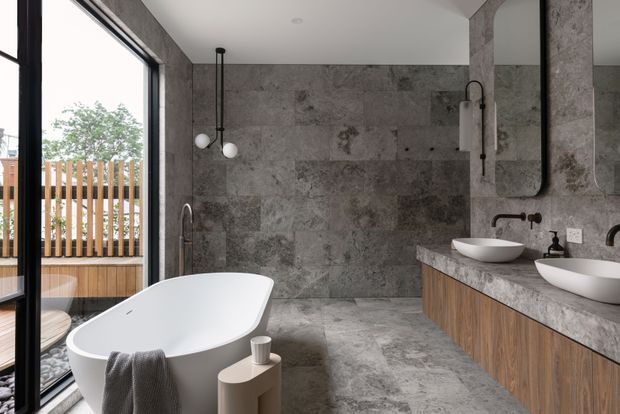 A contemporary bathroom with full-height mottled grey stone-look wall and floor surfaces, a freestanding white bathtub, double vessel basins on a timber vanity, black tapware, and a large frameless mirror. Black-framed glazing opens to a timber deck with vertical screening, creating a strong indoor-outdoor connection.
