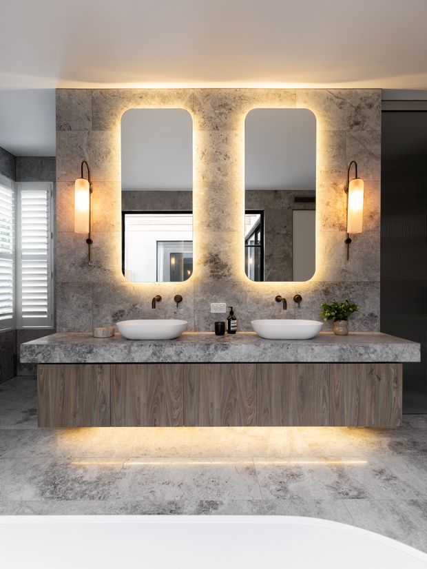 A refined contemporary bathroom featuring a double floating timber-veneer vanity with two white vessel basins, wall-mounted brushed metal taps, and a grey stone-look benchtop and wall finish. Two rounded-rectangle mirrors are backlit against the textured stone wall, complemented by warm vertical sconces. Large grey floor tiles, under-vanity lighting, plantation shutters, and a partial freestanding bath create a calm, luxurious atmosphere.