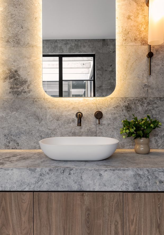 Contemporary bathroom vanity featuring a white vessel basin on a grey veined stone benchtop, with matching large-format grey stone wall tiles and warm backlit rounded-rectangle mirror. Dark bronze or black tapware and wall sconce accents contrast with the timber-look vanity cabinetry below.