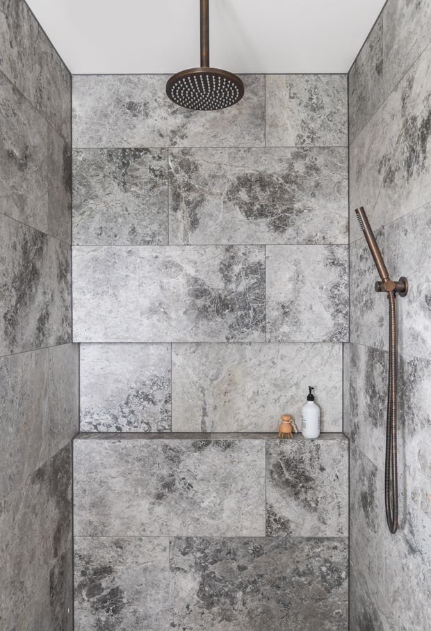 Compact walk-in shower enclosure clad floor-to-ceiling in large-format mottled grey stone-look tiles. The shower features a bronze ceiling-mounted rain shower, matching handheld shower rail and hose, and a built-in tiled niche or bench with small toiletries. The space has a clean, contemporary, monochromatic appearance with strong natural stone variation.