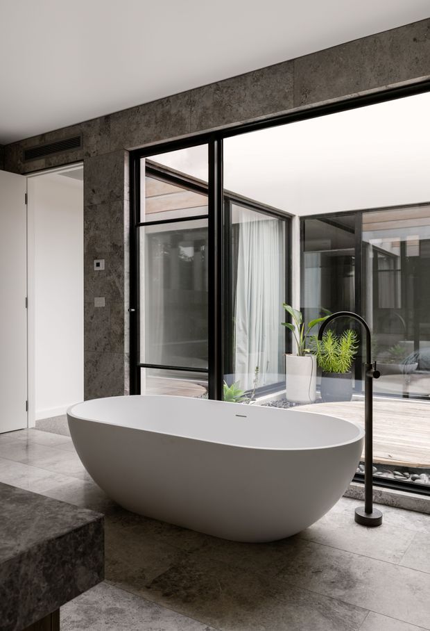 A contemporary bathroom featuring a freestanding white oval bathtub beside a large black-framed glazed opening overlooking an internal courtyard. The room has dark grey stone-look wall and floor finishes, a sculptural black floor-mounted bath filler, and potted plants visible near the glazing. A dark stone-clad platform or bench occupies the foreground.