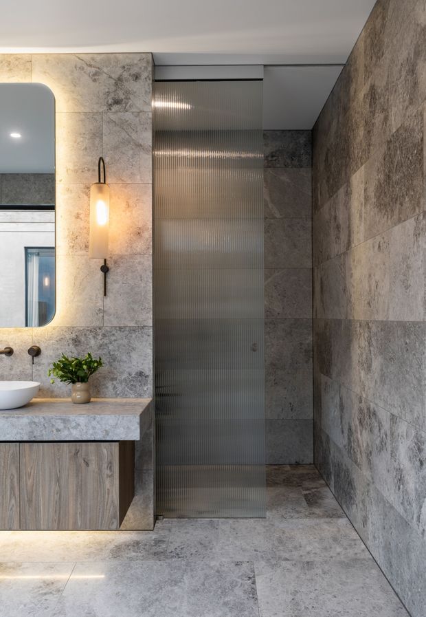 Contemporary bathroom featuring large-format grey stone-look wall and floor tiles, a floating timber vanity with a stone benchtop and vessel basin, rounded-edge mirror, warm wall lighting, and a ribbed translucent glass shower screen.