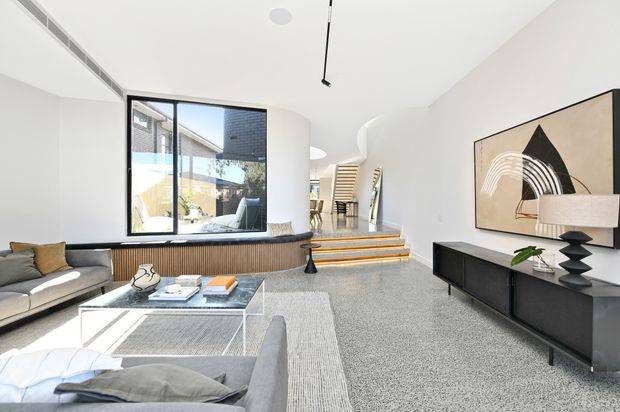 Bright, contemporary open-plan living room with a large grey sectional sofa, glass coffee table, black media console, abstract artwork and sculptural lamp. The room features polished terrazzo flooring, white curved walls and ceilings, extensive black-framed glazing, and a short set of steps leading toward a dining area and staircase. A courtyard or pool area is visible through the glazing.