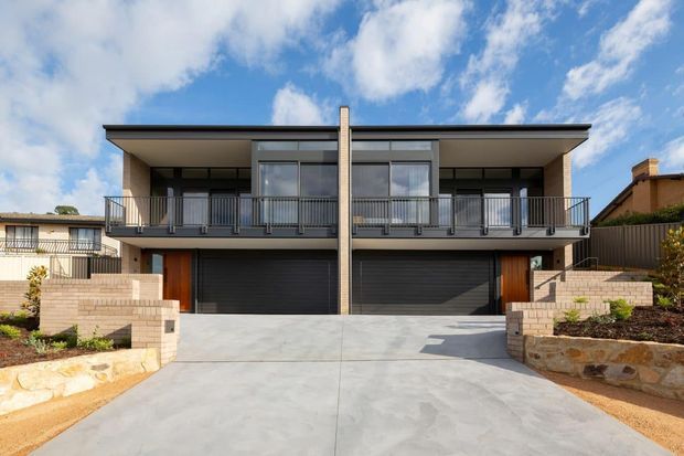 Front exterior view of a contemporary two-storey duplex-style residence with a symmetrical facade, broad upper-level balconies, dark metal garage doors, timber entry doors, exposed brickwork, and a wide concrete driveway. The home is set behind low masonry and stone retaining walls under a bright partly cloudy sky.