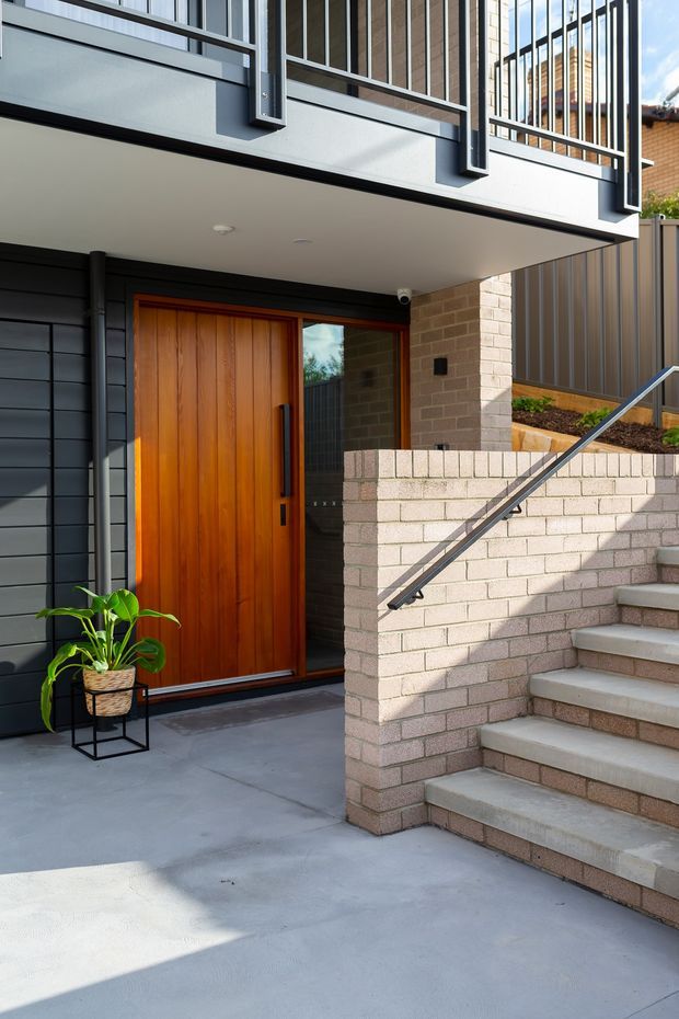 A contemporary residential entry viewed from the exterior, featuring a warm timber front door set within dark horizontal cladding and pale brickwork. A broad concrete stair rises beside a low brick retaining wall with a simple metal handrail, while an upper-level balcony has dark metal vertical balustrades. The entry is sheltered beneath a projecting upper floor and includes a potted plant.