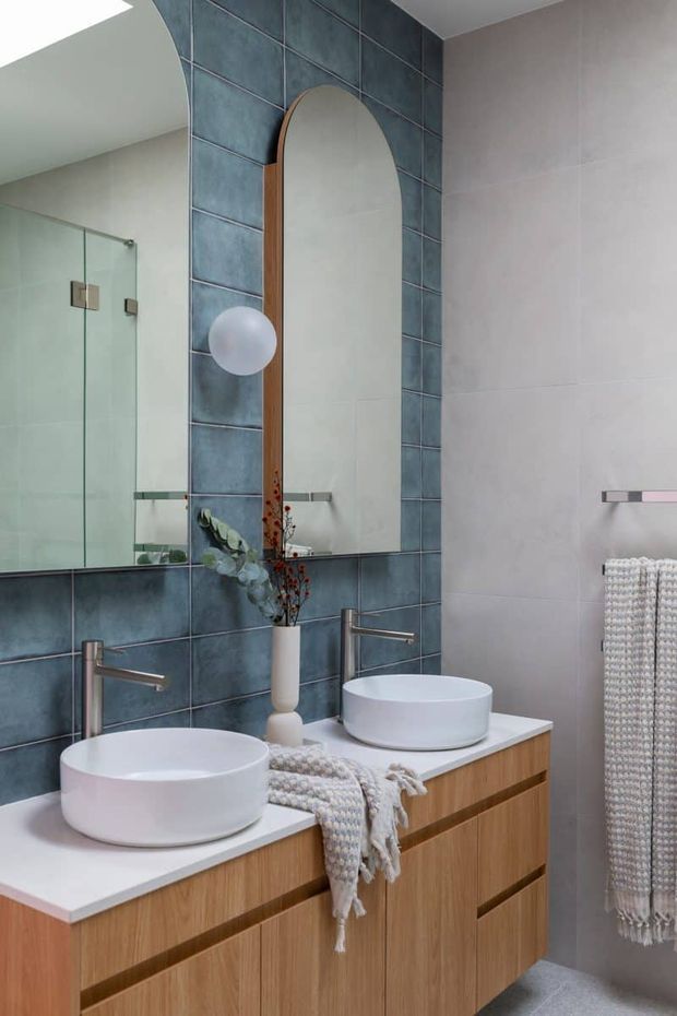 A contemporary bathroom featuring a double vanity with white vessel basins, brushed metal wall-mounted taps, warm timber cabinetry, a white benchtop, and a large blue-grey tiled wall. An arched timber-framed mirror, glass shower enclosure, folded textiles, and soft decorative greenery complete the refined space.