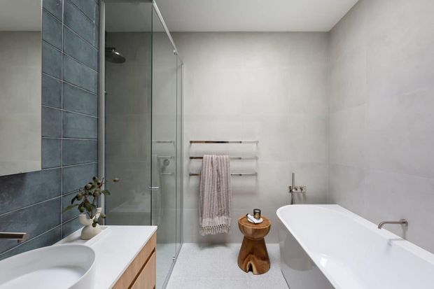 Contemporary bathroom with a freestanding white bathtub, glass-enclosed shower, double vanity, and timber cabinetry. Large pale wall tiles are contrasted by a dark blue-grey tiled feature wall, with warm timber towel rails and a small sculptural stool adding natural detail.