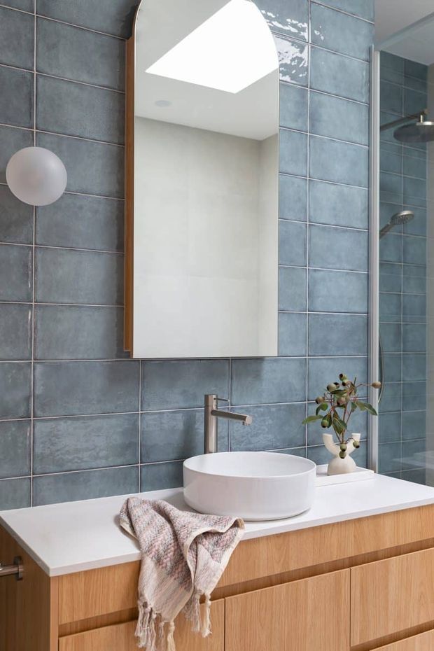 Contemporary bathroom vanity with a white vessel basin, brushed metal tapware, timber-look cabinetry and a white benchtop. Large blue-grey rectangular tiles cover the walls, with a curved-top timber-framed mirror, round wall light and glazed shower enclosure visible at right.