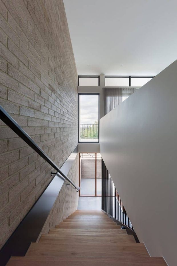 A modern interior stairwell viewed from the upper landing, with broad timber stair treads, exposed pale brick walls, black metal handrails and balustrades, and a tall glazed opening bringing in natural light. The stair rises through a bright, minimalist double-height circulation space.