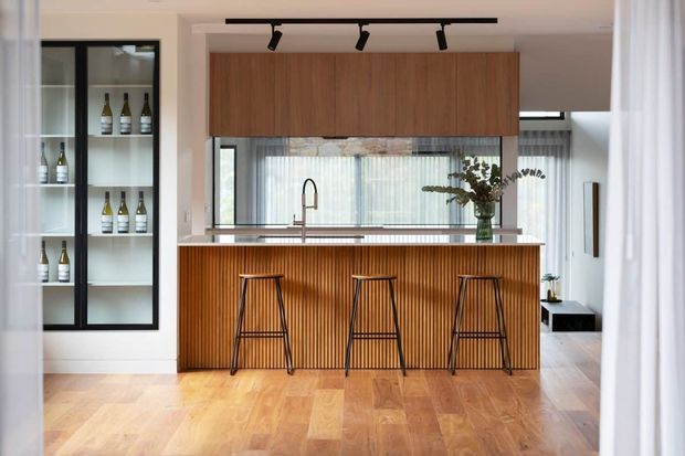 Bright contemporary kitchen centred on a timber island with vertical fluted detailing, a light stone benchtop, integrated timber overhead cabinetry, black-framed glazed wine storage, and three black stools. Timber flooring, sheer curtains, track lighting, and large windows create a warm, refined open-plan setting.