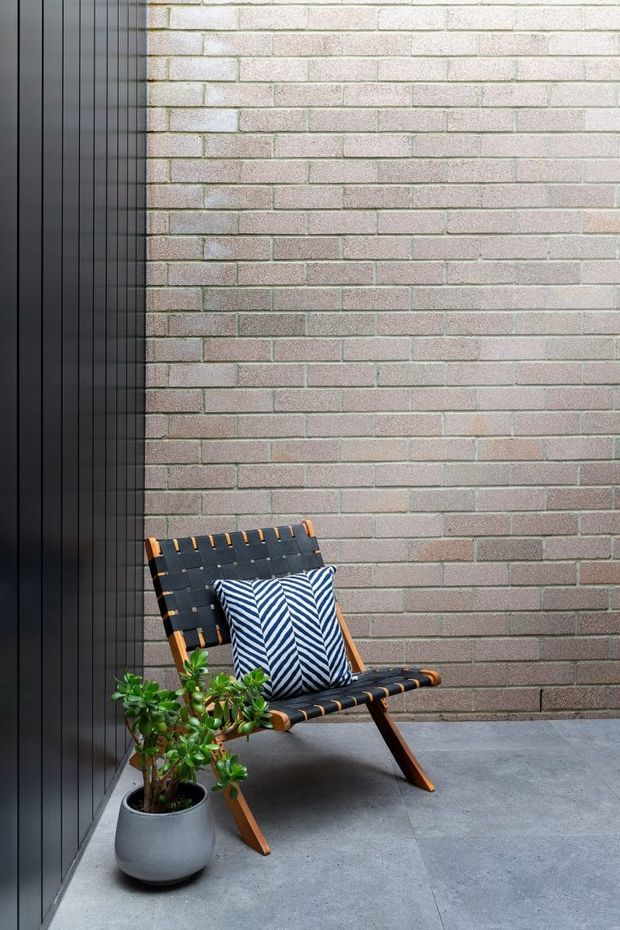 A compact outdoor courtyard or patio corner with a grey brick feature wall, dark vertical metal wall cladding, and large-format grey paving. A black-and-tan sling lounge chair with a blue-and-white patterned cushion sits beside a potted green plant, creating a contemporary urban retreat.